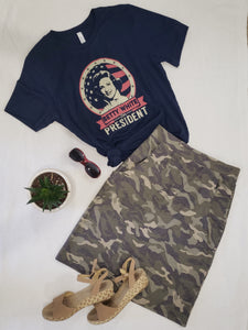 Image of knee length camo printed pencil skirt with pockets, paired with a Vote For Betty campaign graphic tee shirt. Sandels and sunglasses.